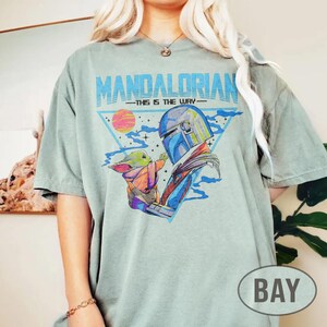 May include: A pale green t-shirt with a graphic of the Mandalorian and Grogu, and the text "MANDALORIAN - THIS IS THE WAY" in blue. The design includes a triangle, a sunset, and a space scene.