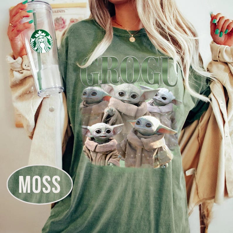 May include: Moss green t-shirt featuring the word "GROGU" above a graphic of five Grogu characters. The shirt has a vintage look. A clear Starbucks tumbler is held in the left hand. The word "MOSS" is in a white oval.