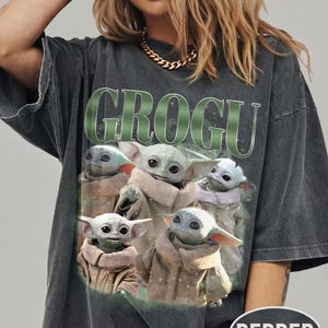 May include: Dark gray t-shirt featuring the word "GROGU" in green block letters above a graphic of Grogu, also known as Baby Yoda, from The Mandalorian. The graphic shows Grogu in various poses. The shirt has short sleeves.