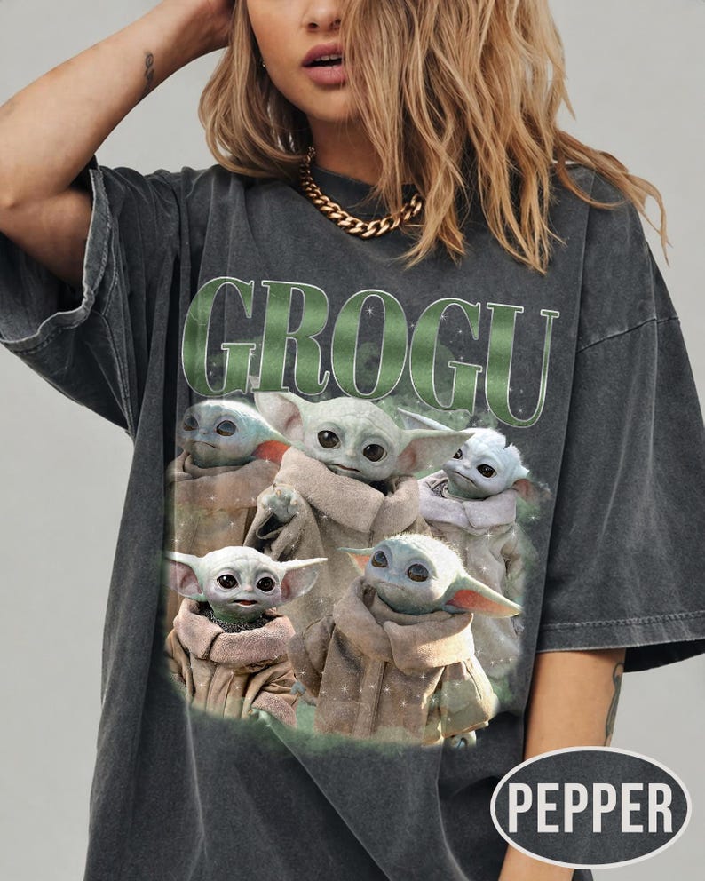 May include: Dark gray t-shirt featuring the word "GROGU" in green block letters above a graphic of Grogu, also known as Baby Yoda, from The Mandalorian. The graphic shows Grogu in various poses. The shirt has short sleeves.