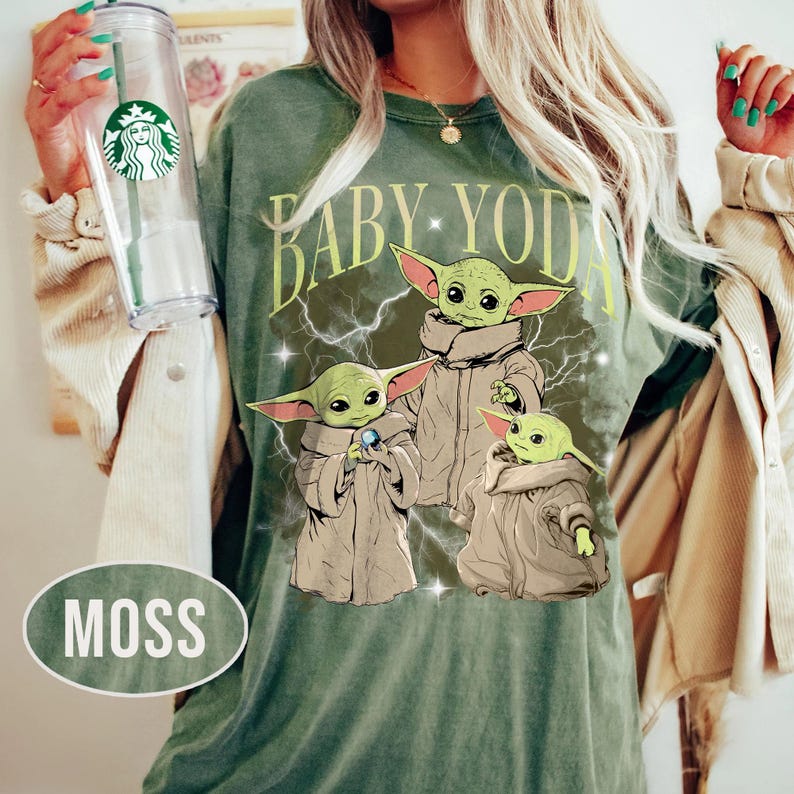 May include: A moss green t-shirt featuring a graphic of three Baby Yoda characters in tan robes surrounded by white lightning bolts and stars. The text BABY YODA is printed in a light yellow font above the characters. A clear plastic cup is visible.