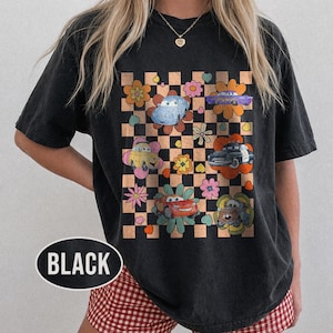 May include: Black t-shirt with a colourful graphic of cartoon cars in flower designs on a chequered background. The shirt has a crew neck and short sleeves. The word "BLACK" is printed on the bottom left of the shirt.