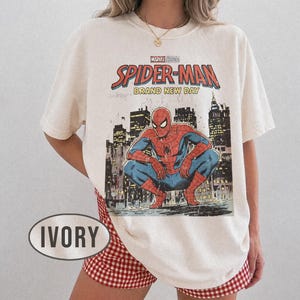 Może przedstawiać: Kremowa koszulka z grafiką Spider-Mana w stylu vintage. Projekt zawiera tekst "Marvel Studios Spider-Man Brand New Day" i ilustrację Spider-Mana w miejskim otoczeniu. Koszulka jest połączona z czerwono-białymi szortami w kratę.
