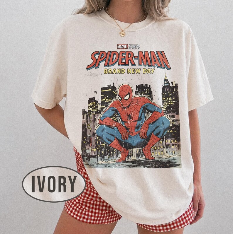 Może przedstawiać: Kremowa koszulka z grafiką Spider-Mana w stylu vintage. Projekt zawiera tekst "Marvel Studios Spider-Man Brand New Day" i ilustrację Spider-Mana w miejskim otoczeniu. Koszulka jest połączona z czerwono-białymi szortami w kratę.