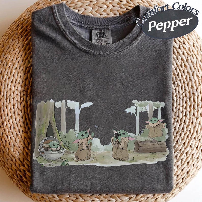 K&ouml;nnte beinhalten: Dunkelgraues Comfort Colors T-Shirt mit einer Cartoon-Illustration einer Figur aus einem beliebten Science-Fiction-Franchise. Die Grafik zeigt die Figur in einer Waldkulisse mit anderen Figuren. Das T-Shirt ist auf einer gewebten Oberfl&auml;che gefaltet.
