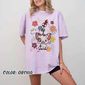 Puede incluir: Camiseta color orqu&iacute;dea con un dise&ntilde;o de un cerdo y un gallo rodeados de flores coloridas. El texto "COLOR: ORCHID" est&aacute; en la parte inferior. La camiseta tiene mangas cortas y un corte holgado.