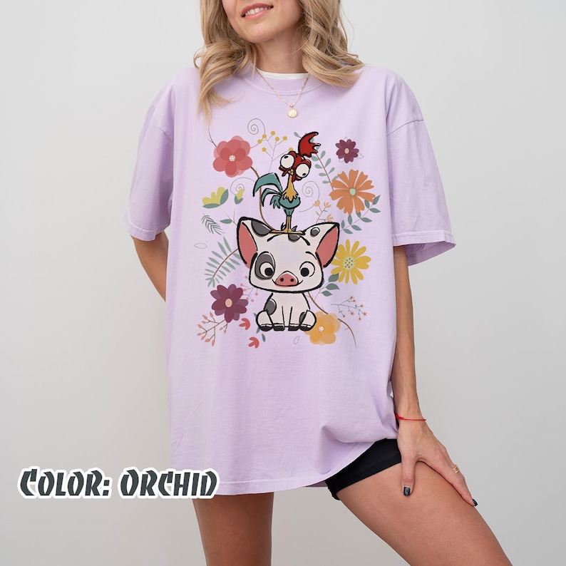 Puede incluir: Camiseta color orqu&iacute;dea con un dise&ntilde;o de un cerdo y un gallo rodeados de flores coloridas. El texto "COLOR: ORCHID" est&aacute; en la parte inferior. La camiseta tiene mangas cortas y un corte holgado.