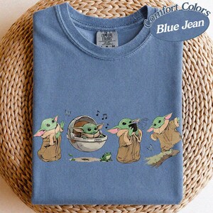 May include: Blue t-shirt featuring Grogu from The Mandalorian. The design shows Grogu in different poses, including one in a floating pod, and the text "Comfort Colors Blue Jean".