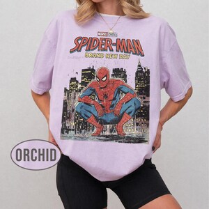 Może przedstawiać: Jasnofioletowa koszulka z grafiką Spider-Mana w kucającej pozie na tle panoramy miasta. Tekst "MARVEL COMICS SPIDER-MAN BRAND NEW DAY" jest wydrukowany w kolorze czerwonym i białym. Koszulka jest w stylu casual.