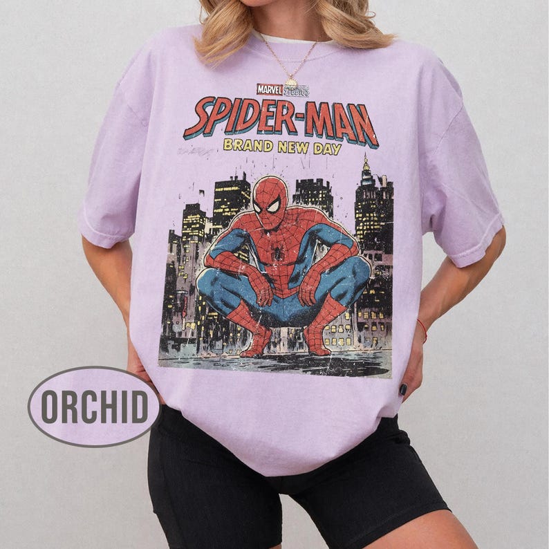 Może przedstawiać: Jasnofioletowa koszulka z grafiką Spider-Mana w kucającej pozie na tle panoramy miasta. Tekst "MARVEL COMICS SPIDER-MAN BRAND NEW DAY" jest wydrukowany w kolorze czerwonym i białym. Koszulka jest w stylu casual.