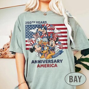 以下が含まれることがあります： アメリカ国旗を背景に、愛国的な衣装を着たディズニーキャラクターのグラフィックが描かれた、薄緑色のTシャツ。「250th Years Anniversary America」の文字が赤と青で書かれています。