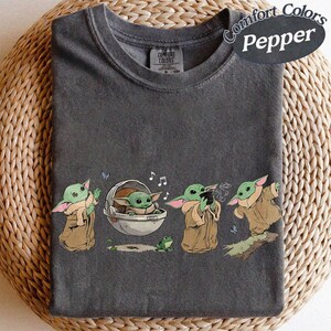 May include: Dark grey t-shirt featuring a cartoon character from a popular science fiction series. The design shows the character in different poses, with musical notes and a frog. The shirt is labelled "Comfort Colors Pepper".