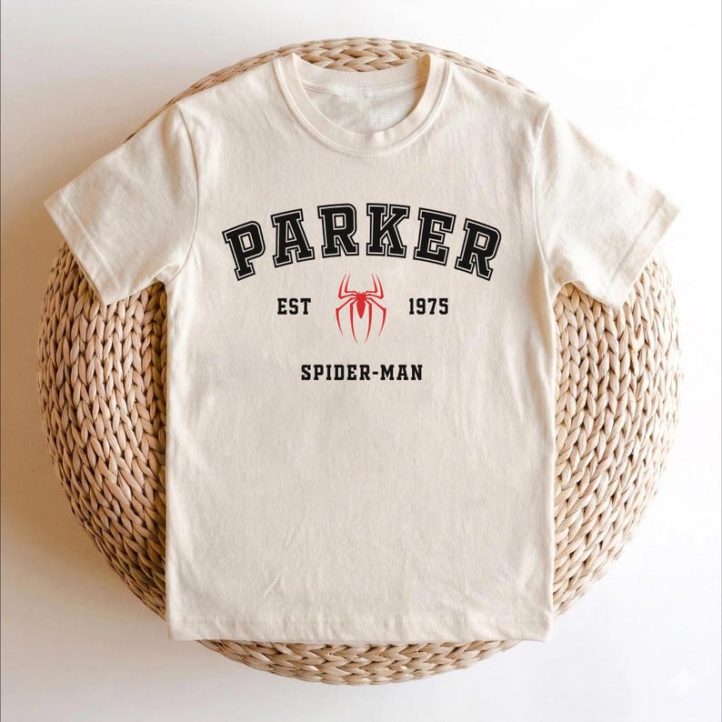 May include: Cream-colored t-shirt with the word "PARKER" in black arched lettering. Below the word "PARKER" is a red spider emblem, with the text "EST 1975" on either side. The words "SPIDER-MAN" are printed below the emblem.