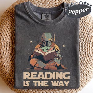 May include: Pepper-coloured t-shirt with a graphic of a Mandalorian character reading a book with Grogu. The design includes the text "READING IS THE WAY" in tan. The shirt is made of a soft material.