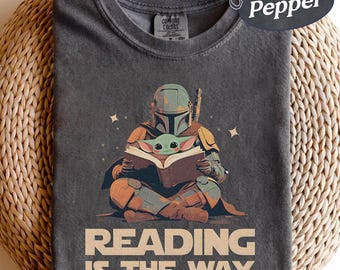 Op de afbeelding: Pepperkleurig t-shirt met een afbeelding van een Mandalorian personage dat een boek leest met Grogu. Het ontwerp bevat de tekst "READING IS THE WAY" in beige. Het shirt is gemaakt van een zacht materiaal.