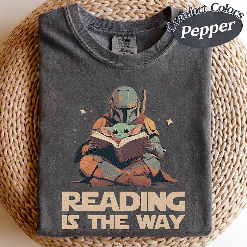 May include: Pepper-coloured t-shirt with a graphic of a Mandalorian character reading a book with Grogu. The design includes the text "READING IS THE WAY" in tan. The shirt is made of a soft material.