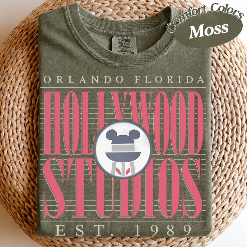 May include: A moss green t-shirt with the words "Orlando Florida Hollywood Studios" in white and pink. The shirt features a Mickey Mouse head logo and the text "EST. 1989". The shirt is folded on a woven surface.