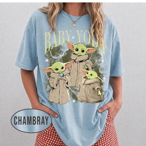 May include: A light blue short-sleeve t-shirt featuring a graphic print of three green creatures in tan robes. The text BABY YODA is printed in yellow above the characters, which are set against a dark, lightning-filled background. The word CHAMBRAY appears in a gray oval at the bottom left.