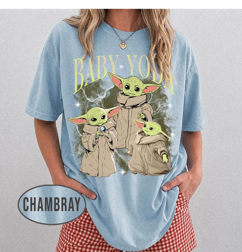 May include: A light blue short-sleeve t-shirt featuring a graphic print of three green creatures in tan robes. The text BABY YODA is printed in yellow above the characters, which are set against a dark, lightning-filled background. The word CHAMBRAY appears in a gray oval at the bottom left.