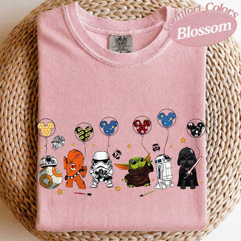 May include: A pink Comfort Colors Blossom t-shirt featuring Star Wars characters and Mickey Mouse-themed balloons. The design includes BB-8, Chewbacca, a Stormtrooper, Grogu, R2-D2, and Darth Vader. The shirt has a crew neck.