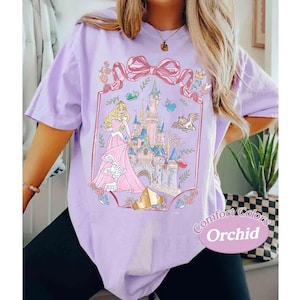 May include: A lilac Comfort Colors t-shirt featuring a princess and castle graphic. The graphic includes a pink bow, castle, and the text "Comfort Colors Orchid". The princess is wearing a pink gown.