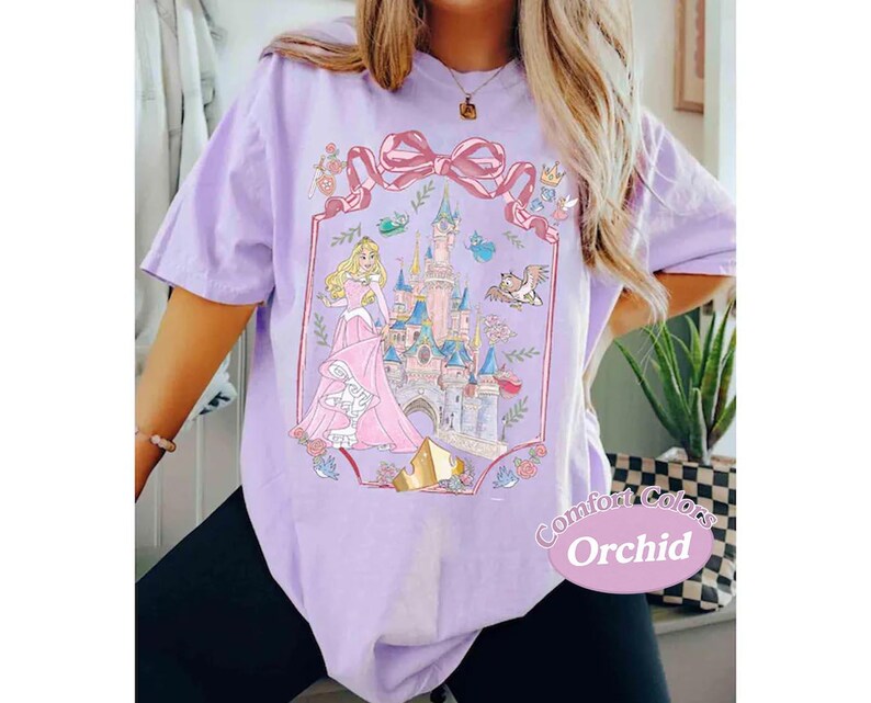 May include: A lilac Comfort Colors t-shirt featuring a princess and castle graphic. The graphic includes a pink bow, castle, and the text "Comfort Colors Orchid". The princess is wearing a pink gown.