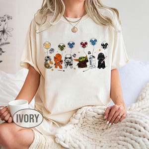 May include: Ivory-colored t-shirt featuring a line of Star Wars characters holding Mickey Mouse-themed balloons. Characters include BB-8, Chewbacca, a Stormtrooper, Grogu, R2-D2, and Darth Vader. The word "IVORY" is printed on the front of the shirt.