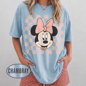 May include: Light blue t-shirt featuring a Minnie Mouse graphic with a pink bow and a checkered background. The shirt has short sleeves and a relaxed fit. The word "CHAMBRAY" is printed on the bottom left of the shirt.