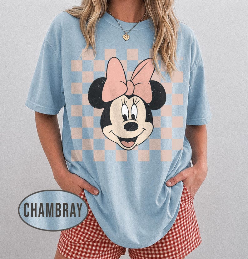 May include: Light blue t-shirt featuring a Minnie Mouse graphic with a pink bow and a checkered background. The shirt has short sleeves and a relaxed fit. The word "CHAMBRAY" is printed on the bottom left of the shirt.