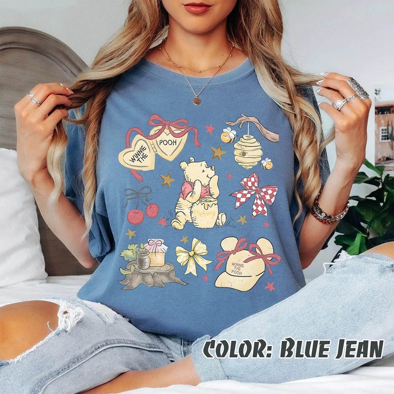 May include: Blue jean coloured t-shirt with a Winnie the Pooh graphic. The design includes illustrations of Pooh Bear, honey pots, bows, hearts, cherries, and a beehive. The text "Winnie the Pooh" is also present.