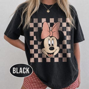 May include: Black t-shirt featuring a Minnie Mouse graphic with a pink bow and a checkered background. The word "BLACK" is printed on the bottom left of the shirt in a white oval.