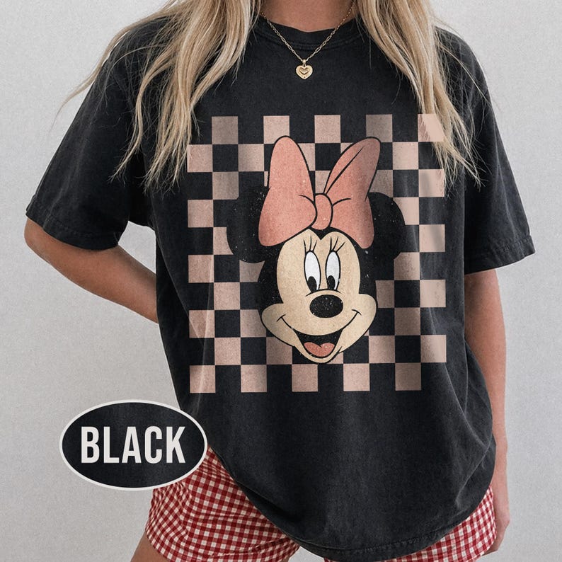 May include: Black t-shirt featuring a Minnie Mouse graphic with a pink bow and a checkered background. The word "BLACK" is printed on the bottom left of the shirt in a white oval.