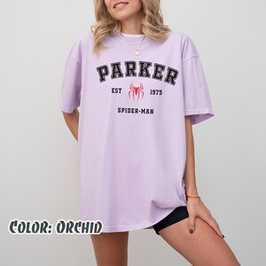 May include: Orchid-colored t-shirt with the word "PARKER" in black block letters, "EST 1975" and "SPIDER-MAN" printed below. A red spider graphic is centered between "EST" and "1975". The text "COLOR: ORCHID" is at the bottom.