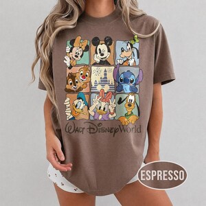Op de afbeelding: Espresso-kleurig T-shirt met een raster van Disney-personages, waaronder Mickey Mouse, Minnie Mouse, Goofy, Donald Duck, Katrien Duck, Knabbel en Babbel, Stitch en Pluto. De woorden "Walt Disney World" staan eronder.