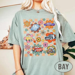 May include: Sage green t-shirt with a colourful graphic of cartoon cars and flowers. The design includes a checkered background. The word "BAY" is printed on the bottom right.