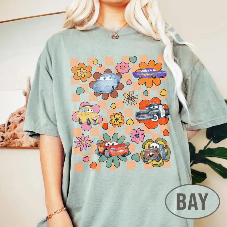 May include: Sage green t-shirt with a colourful graphic of cartoon cars and flowers. The design includes a checkered background. The word "BAY" is printed on the bottom right.