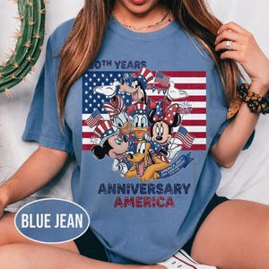 以下が含まれることがあります： アメリカ国旗の前にディズニーキャラクターが描かれた、ブルーデニムカラーのTシャツ。ミッキーマウス、ミニーマウス、ドナルドダック、プルートが含まれており、「60th Years Anniversary America」の文字が書かれています。