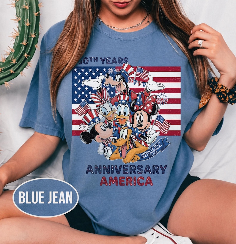 以下が含まれることがあります： アメリカ国旗の前にディズニーキャラクターが描かれた、ブルーデニムカラーのTシャツ。ミッキーマウス、ミニーマウス、ドナルドダック、プルートが含まれており、「60th Years Anniversary America」の文字が書かれています。