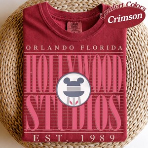May include: Crimson Comfort Colors t-shirt with pink text reading "Orlando Florida Hollywood Studios". Includes a Mickey Mouse icon and "EST. 1989". The shirt is folded on a woven surface.