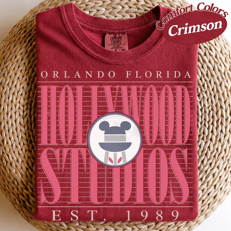 May include: Crimson Comfort Colors t-shirt with pink text reading "Orlando Florida Hollywood Studios". Includes a Mickey Mouse icon and "EST. 1989". The shirt is folded on a woven surface.