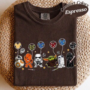 May include: Espresso-colored t-shirt featuring a line of Star Wars characters holding Mickey Mouse-shaped balloons. Characters include BB-8, Chewbacca, a Stormtrooper, Grogu, R2-D2, and Darth Vader. The shirt has a Comfort Colors label.