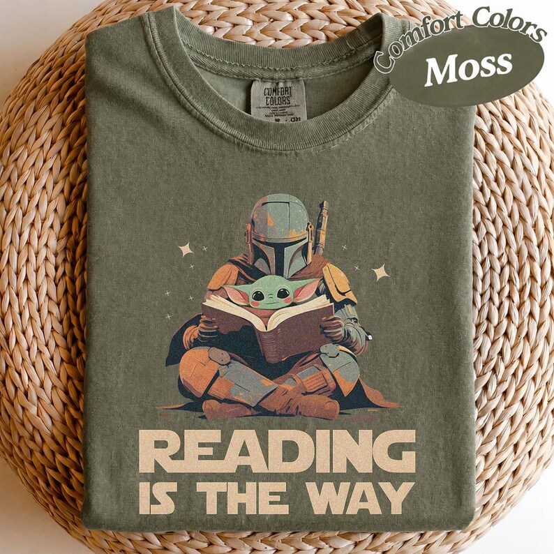 May include: Moss green t-shirt with a graphic of the Mandalorian and Grogu reading a book. The text "READING IS THE WAY" is printed below the illustration. The shirt is a Comfort Colors brand.