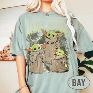 May include: A sage green short-sleeve t-shirt featuring a graphic print of three green creatures with large ears and tan robes. The text "BABY YODA" is printed in a light yellow font above the characters, set against a dark, cloudy background.
