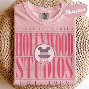 May include: A pale pink t-shirt with the words "Orlando Florida Hollywood Studios" in a retro font. The shirt features a Mickey Mouse head logo and the text "EST. 1989". The shirt is labelled "Comfort Colors Blossom".