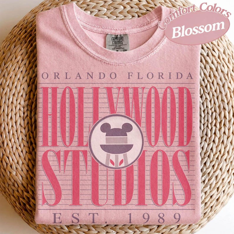 May include: A pale pink t-shirt with the words "Orlando Florida Hollywood Studios" in a retro font. The shirt features a Mickey Mouse head logo and the text "EST. 1989". The shirt is labelled "Comfort Colors Blossom".