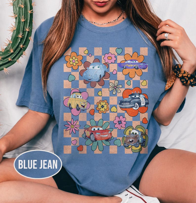 May include: Blue jean t-shirt with a vibrant graphic of cartoon cars and flowers on a checkered pattern. The tee has a crew neck and short sleeves. The text "BLUE JEAN" is printed on the bottom left of the shirt.