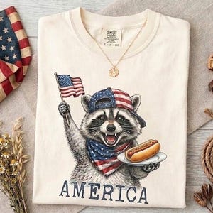Könnte beinhalten: Ein cremefarbenes T-Shirt mit einer Grafik eines Waschbären, der eine Baseballkappe und ein Bandana im Sternenbanner-Design trägt und eine kleine amerikanische Flagge sowie einen Hotdog auf einem Teller hält. Darunter steht AMERICA in Blau.