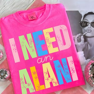 May include: Bright pink crew neck sweatshirt with the text "I NEED an ALANI" in colorful, distressed lettering. The sweatshirt is a vibrant, eye-catching piece of apparel.