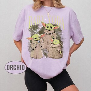 May include: A light purple short-sleeve t-shirt featuring a graphic print of three green creatures in tan robes. The text BABY YODA is printed in yellow above the characters, set against a dark, cloudy background with lightning effects.