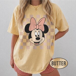 May include: Butter-colored t-shirt featuring a Minnie Mouse graphic with a pink bow and a checkered background. The word "BUTTER" is printed on the bottom right. The shirt has short sleeves and a relaxed fit.